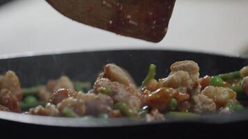 The chef of Asian cuisine combines Chinese chicken with a golden crust in a sweet and sour sauce, along with green peas and green beans, in a frying pan using a wooden spoon. Extreme close-up. Slow motion. 4k video