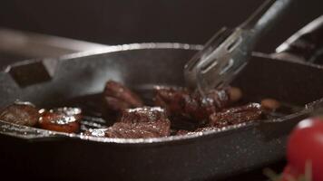 A professional chef is roasting pieces of beef in a black pan on an induction hob. In the foreground are tomatoes, and the background is black. Close up. Slow motion. 4k video