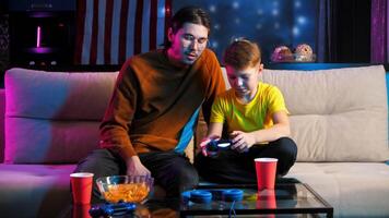 dans le soir, le plus âgée frère est assis sur le canapé et enseigne le plus jeune frère Comment à utilisation le manette pour le Jeu console. milieu tir. 4k video