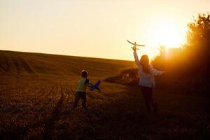 Running boy and girl holding two yellow and blue airplanes toy in the field during summer sunset. Kids dreams of flying and aviation. photo