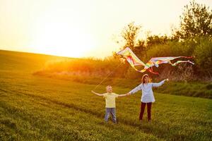 Smiling girl and brother boy standing and holding hands with flying colorful kites on the high grass meadow. Happy childhood moments photo