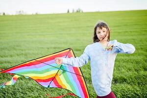 pequeño contento niña corriendo con Kate en manos en verde trigo campo. grande de colores arco iris cometa con largo cola. foto