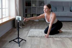 mujer sentado en el piso en yoga pantalones grabación un en línea aptitud con su teléfono y trípode, sonriente a el cámara en un vivo habitación ajuste foto