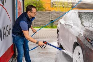 Young man washes his car at a self-service car wash using a hose with pressurized water and foam. High quality photo