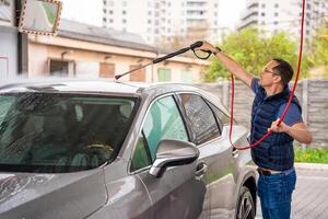 Young man washes his car at a self-service car wash using a hose with pressurized water and foam. High quality photo