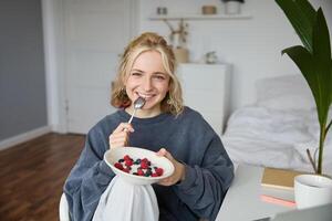 Portrait of beautiful young woman in a room, eating breakfast, holding bowl with dessert and a spoon, smiling at camera, recording lifestyle vlog photo