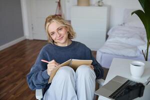 retrato de sonriente linda mujer, estilo de vida bloguero, se sienta en su habitación con diario diario o planificador, registros en digital cámara, crea contenido para social medios de comunicación acerca de diario rutina foto