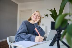 retrato de joven mujer, estilo de vida bloguero, grabación de sí misma, haciendo notas, escritura en diario, sentado en frente de ordenador portátil en un habitación y estudiando foto