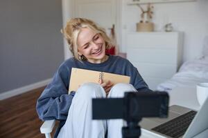 Social media and lifestyle content concept. Young happy smiling woman, sits in her room with notebook, talks at digital camera, having conversation with followers, does lifestyle vlog photo