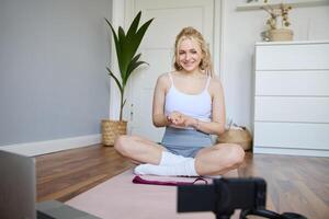 Portrait of young charismatic fitness trainer, girl blogger records on digital camera, talks about health and workout, doing exercises on rubber mat in a room at home photo