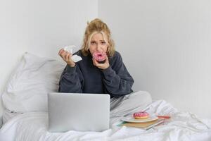 Portrait of sad woman crying, eating doughnut, wiping tears off, looking at something upsetting on laptop screen, sitting on a bed photo