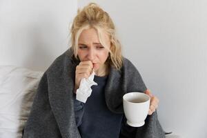 Portrait of woman catching a cold, staying home sick, drinking tea, sneezing in napkin, blowing nose, has covid symptoms photo