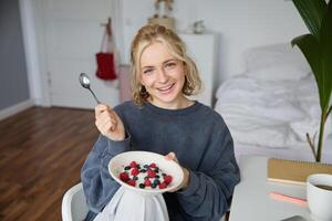 retrato de sonriente rubio mujer, comiendo desayuno, participación cuenco y cuchara, sentado en dormitorio, mirando contento a cámara foto