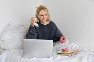 Portrait of woman sitting on a bed with laptop, eating doughnut and crying from sad movie scene photo