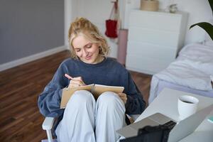 retrato de joven mujer escritura en diario, haciendo notas en computadora portátil, sentado en silla en un habitación en frente de computadora portátil, estudiante haciendo deberes foto