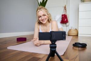 Portrait of young woman, sports vlogger, fitness instructor recording of herself showing workout exercises, using digital camera, lying on yoga rubber mat photo