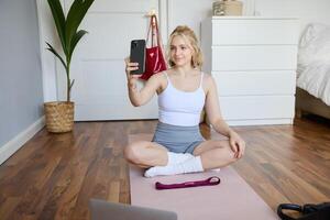 Portrait of stylish young woman, sitting on rubber mat on floor at home, taking selfie while doing workout, yoga session, posting photo on social media