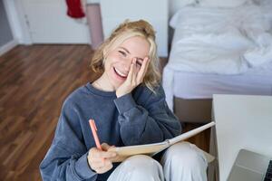 Portrait of happy, young blond woman, sitting in bedroom with notebook and pen, laughing and smiling, writing in journal, doing homework and smiling photo
