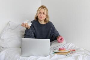 Portrait of disappointed, depressed young woman, crying, sitting on bed with laptop, eating comfort food, holding doughnut and wiping tears off photo
