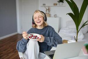 Portrait of happy young blond woman, sitting in a room, watching movie on laptop and eating healthy breakfast, drinking tea, resting on weekend photo