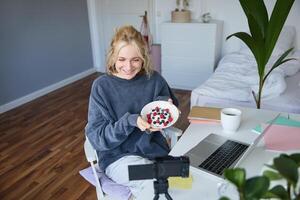 Portrait of cute young girl vlogger, showing her breakfast on camera, recording vlog in her bedroom, holding dessert, eating photo