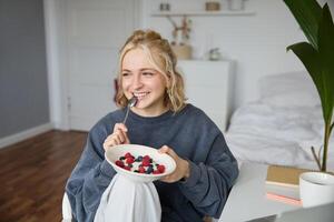 Close up portrait of smiling, cute blond woman, eating healthy lunch in her room, holding bowl with berries, biting spoon, looking happy photo