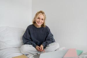 retrato de joven hermosa mujer sentado en cama con ordenador portátil y cuadernos, trabajando desde hogar, trabajo independiente hembra estudiante estudiando en línea, charlas, conecta a lección remotamente foto