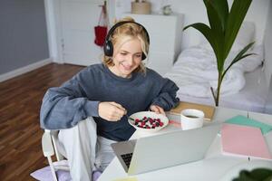 Portrait of happy young blond woman, sitting in a room, watching movie on laptop and eating healthy breakfast, drinking tea, resting on weekend photo