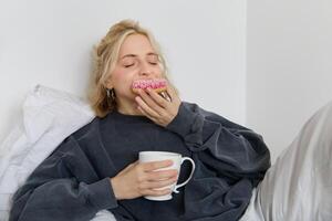 Close up of young blond woman lying in bed with cup of tea, biting delicious doughnut with pleasure and satisfied face photo