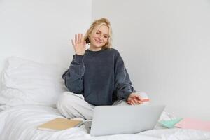 Portrait of beautiful, smiling young woman sitting on bed, waves her hand at laptop camera, says hello to friend on chat app, studies at home on remote, connects to online course photo