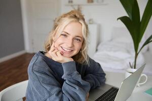 Close up portrait of young smiling female model, sits in a room with laptop, works from home, does online course, e-learning photo