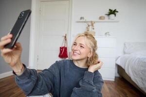 retrato de joven elegante niña se sienta en dormitorio piso, toma selfies en su teléfono inteligente, posando para foto en social medios de comunicación aplicación, sonriente y mirando contento a cámara