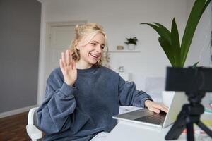 Portrait of young social media content creator, woman sits in room with laptop and digital camera, records lifestyle vlog, says hello, chats to someone online via computer photo