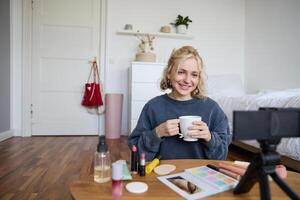 Portrait of beautiful social media beauty blogger, sitting in front of digital camera on floor in bedroom, drinking tea and chatting, talking to followers photo