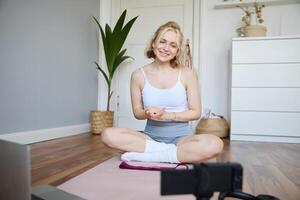 Young female athlete, fitness instructor woman sits on floor rubber mat, recording on digital camera, showing how to workout, explaining exercises photo