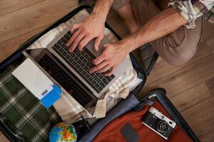 Top view of opened suitcase with clothes, male hands using laptop, booking hotel for vacation, check-in on flight for holiday, packed camera to shoot pictures photo