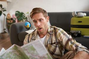 Portrait of man, tourist with concerned face, sits with suitcase, looks at travel map, plans the route photo