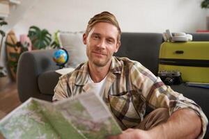 Close up portrait of smiling handsome man, boyfriend plans a holiday, holds travel map, sits near packed suitcase photo