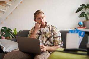 Portrait of man with concerned face, looks aside with worried expression, sits on sofa with laptop, has two tickets on plane and suitcase photo