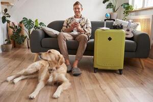 retrato de feliz, hermoso joven hombre con maleta, sentado con su perro en vivo habitación, preparado a Vamos en vacaciones, yendo en fiesta foto