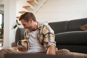 retrato de contento joven hombre, abrazando y jugando con su perro, sentado en piso con sofá, mirando a dorado perdiguero foto