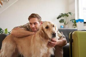 Portrait of young man hugging his dog, going on vacation to pet-friendly hotel, packed suitcase, sits in living room photo