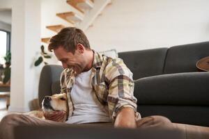 Happy young smiling guy in living room, sitting on floor, hugging his pet, playing with dog photo