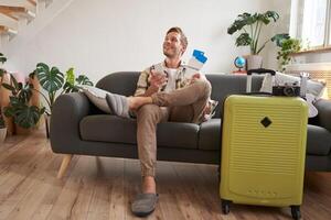 Handsome young man sits on sofa with suitcase, holds smartphone, books hostel or holiday vacation, chats on mobile phone photo