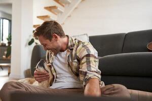 Image of happy, smiling young man playing with his dog, sitting in living room on sofa, working with laptop photo