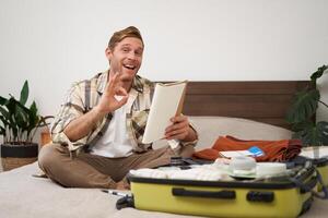 Image of cheerful man shows okay sign, packs clothes in suitcase, goes on holiday or business trip, holds notebook with list of items to take with him photo