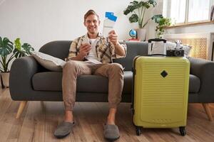 Portrait of happy man checked in online on smartphone, showing tickets in his hand, sitting near suitcase, going on vacation, preparing for holiday photo