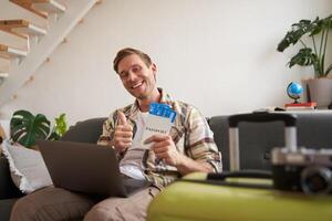 Portrait of happy handsome guy, shows his flight tickets, booked a trip for summer holiday, sits with laptop in living room, smiles at camera photo