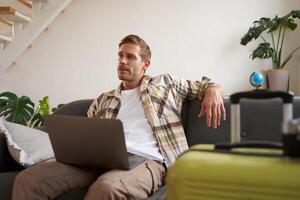 Portrait of happy young man, tourist booking his holiday with travel agency, looking at laptop, packed suitcase to go on vacation photo