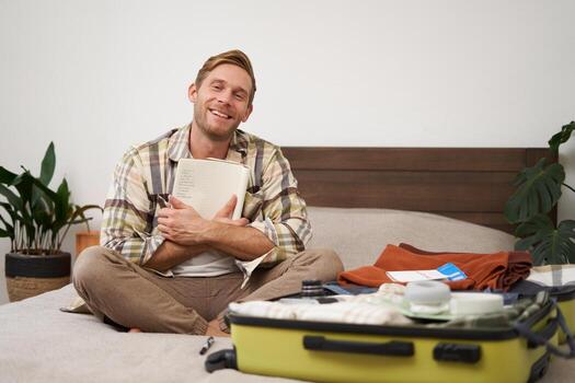 Portrait of charismatic, handsome man, tourist looking at notebook, getting ready for journey, packing suitcase, checking list of items on journal photo